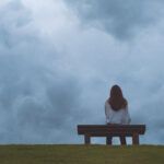 a woman sitting on a bench in front of a cloudy sky.