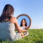 a woman sitting in the grass holding a circular mirror.