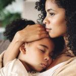 A woman with curly hair holds a sleeping child close to her chest, both seated. The woman has a thoughtful expression, and the background includes a blurred indoor setting.