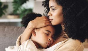 A woman with curly hair holds a sleeping child close to her chest, both seated. The woman has a thoughtful expression, and the background includes a blurred indoor setting.