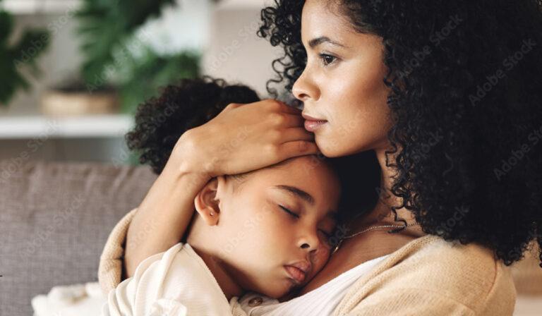 A woman with curly hair holds a sleeping child close to her chest, both seated. The woman has a thoughtful expression, and the background includes a blurred indoor setting.