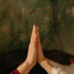 Two people sit facing each other with their hands pressed together, palms touching, in an authentic therapy session against a blurred green background with foliage patterns.