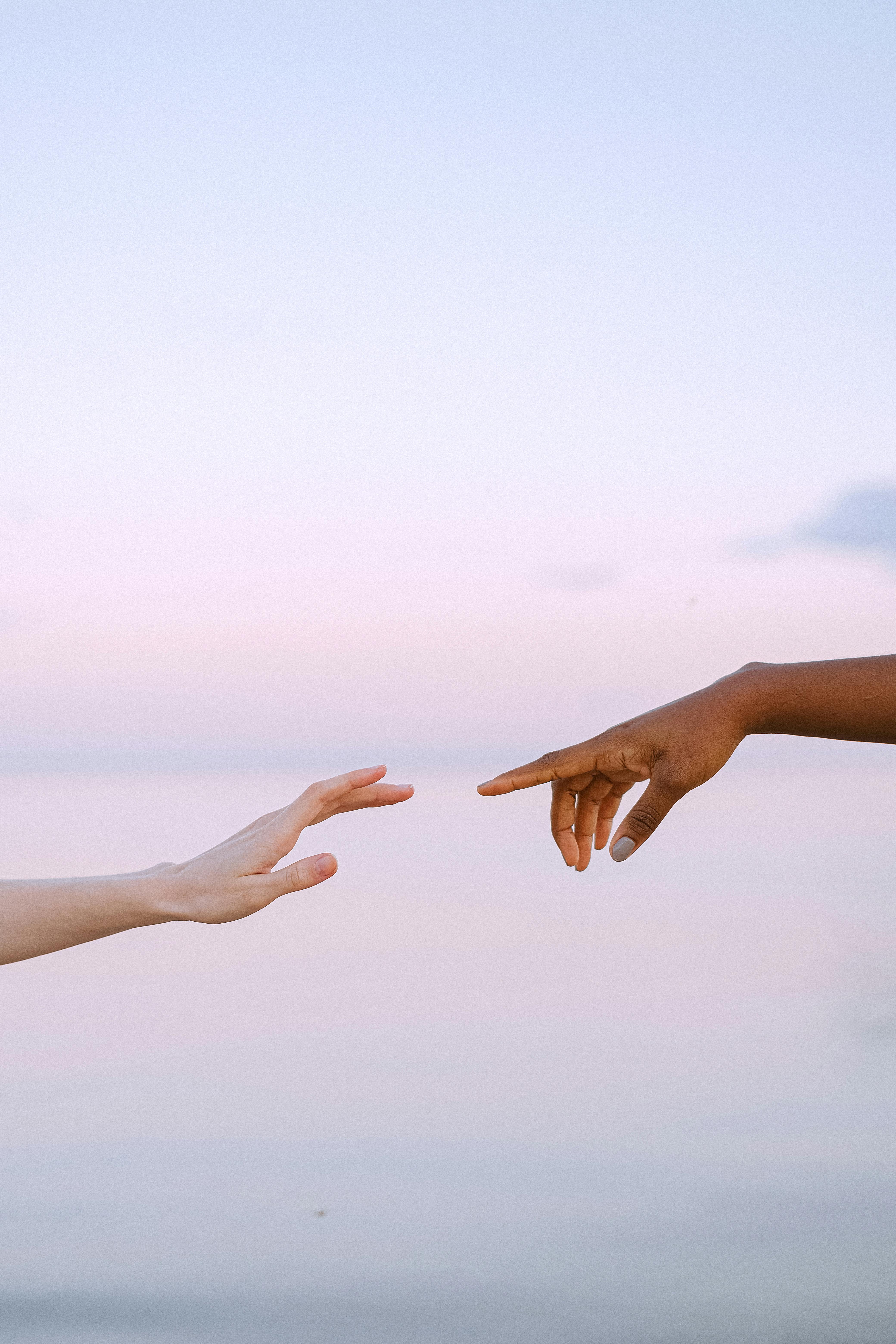 Two hands, one with lighter skin and one with darker skin, reach toward each other against a pastel sky background.
