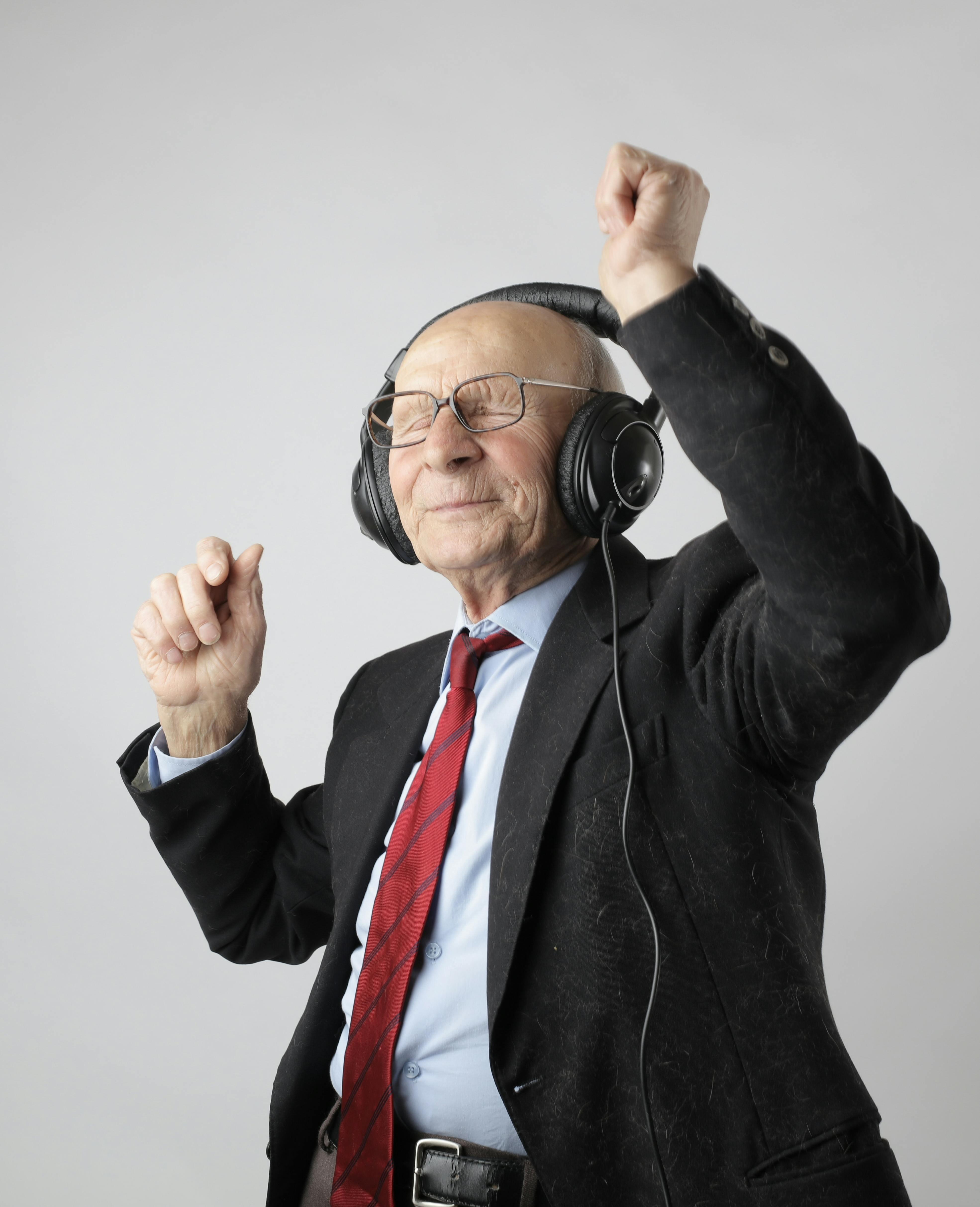 Elderly man in a suit and red tie wearing headphones, smiling with eyes closed, dancing with one arm raised against a plain background.