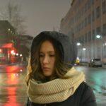 A young woman wearing a hooded coat and knit scarf stands on a wet city street at night, with buildings and streetlights in the background.