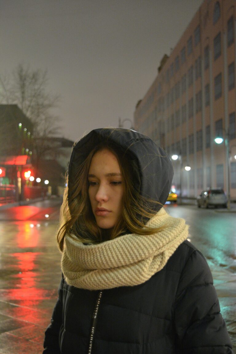A young woman wearing a hooded coat and knit scarf stands on a wet city street at night, with buildings and streetlights in the background.